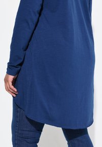 Langärmlige Tunika aus tiefblauem Stoff mit abgerundetem Saum, der eine glatte Textur und enganliegende Ärmel aufweist, kombiniert mit dunkler Denim.