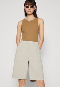 Barna bordázott ujjatlan felső bézs szabott rövidnadrággal párosítva. A rövidnadrág sima textúrájú és redőzött részletekkel rendelkezik, amely struktúrált megjelenést kölcsönöz.