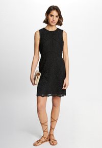 Zwarte kanten jurk met een getailleerd silhouet, mouwloos ontwerp, bloemenpatroon en scalloped zoom. Gepaard met tan gladiatorsandalen en een clutch.