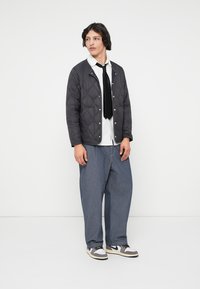 Ung mann står, iført en svart quiltet jakke, hvit skjorte, svart slips, løse blå bukser og hvit-grå joggesko foran en ensfarget hvit bakgrunn.