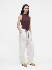 Femme debout, mains dans les poches, portant un top marron sans manches, un pantalon blanc ample avec cordon et des sandales jaunes sur fond blanc.