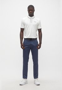 Άνδρας στέκεται φορώντας λευκό πόλο με μαύρη λεπτομέρεια, μπλε σκούρο παντελόνι, μαύρη ζώνη και λευκά αθλητικά παπούτσια, με φόντο ένα απλό λευκό τοίχο.