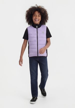 Χαμογελαστό παιδί με σγουρά μαλλιά φοράει γιλέκο από λιλά καπιτονέ ύφασμα πάνω από μαύρο μπλουζάκι με κοντά μανίκια, σκούρο τζιν και μαύρα αθλητικά παπούτσια, περπατώντας προς τα εμπρός.