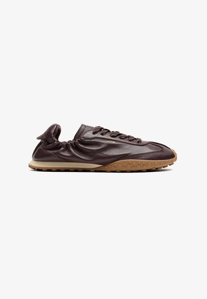 Donkerbruine leren sneaker met een gestructureerde beige rubberen zool en elastische hiel, weergegeven in zijprofiel op een witte achtergrond.