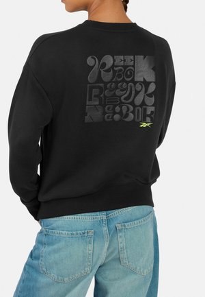 Person trägt schwarzen Sweatshirt mit stilisiertem schwarzem Reebok-Schriftzug und kleinem gelbem Reebok-Logo auf dem Rücken, kombiniert mit zweifarbigen blauen Jeans.