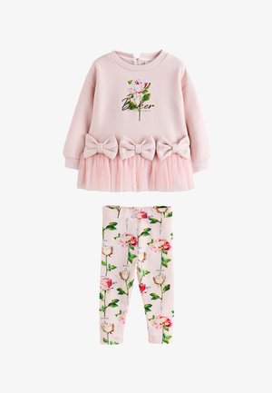 Młoda dziewczyna w różowej bluzie z kwiatowym nadrukiem i tiulowej spódniczce, dopasowanych legginsach w kwiaty oraz białych trampkach stoi na szarej podłodze na tle białej ściany.