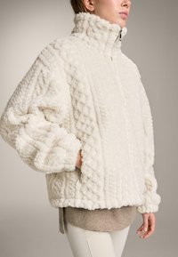 Bílá nadýchaná zipová bunda s vysokým límcem, strukturovaným vzorem a žebrovaným lemem. Rukávy jsou oversized a mírně řasené.