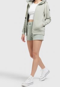 Donna che indossa una felpa verde pallido con zip e pantaloni corti abbinati, un top corto bianco e sneakers bianche alte, in piedi contro uno sfondo neutro.