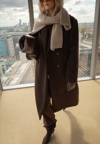 Brązowy wełniany płaszcz na ciemnej stylizacji, szary dziany szal, pikowana brązowa torebka i brązowe rękawiczki; miejska sceneria z panoramą miasta.