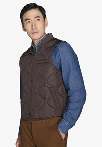 Giubbotto imbottito marrone con colletto alto e zip frontale; abbinato a una camicia in denim blu e pantaloni marroni. La superficie sembra liscia e leggera.