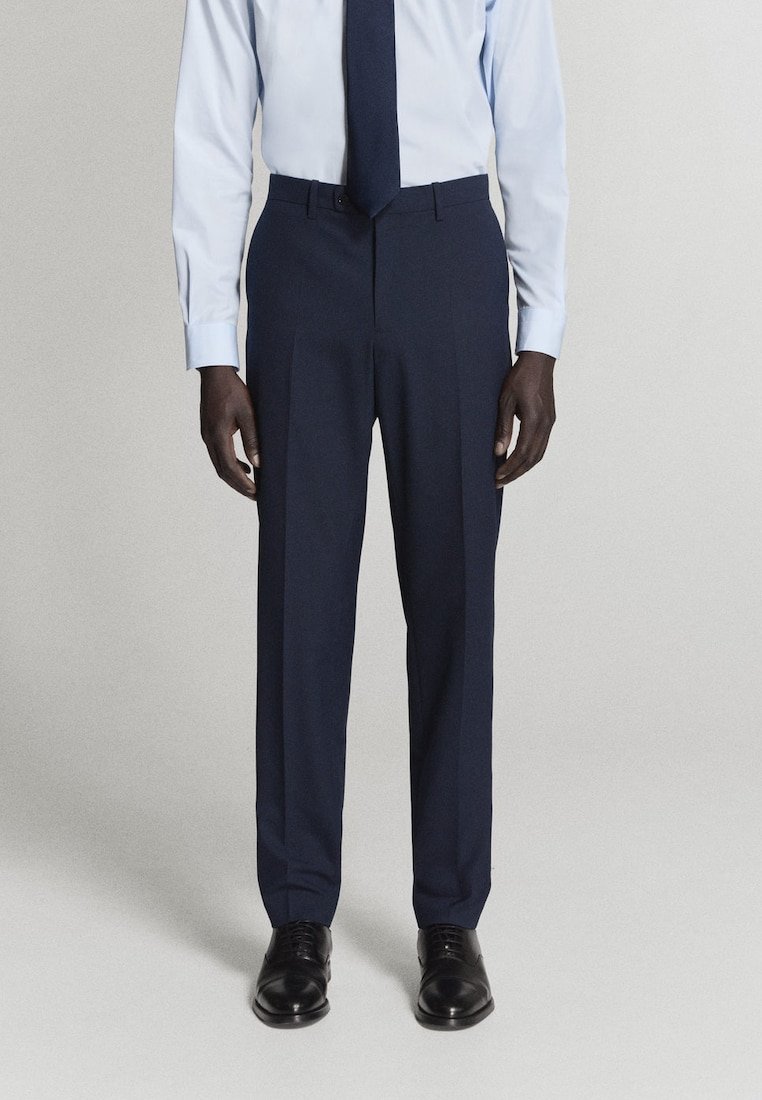 Homme portant un pantalon de costume bleu marine, une chemise bleu clair, une cravate bleu marine et des chaussures habillées noires, debout devant un fond clair uni.