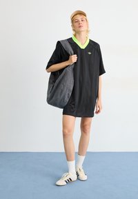 Svart sportklänning med en neongrön V-ringning, i kombination med en grå tygväska och vita sneakers. Mot en ljusblå bakgrund.