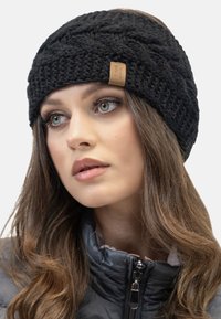 Bandeau en tricot noir avec un motif texturé, arborant une étiquette en cuir cousue. Porté sur des cheveux ondulés de longueur moyenne.