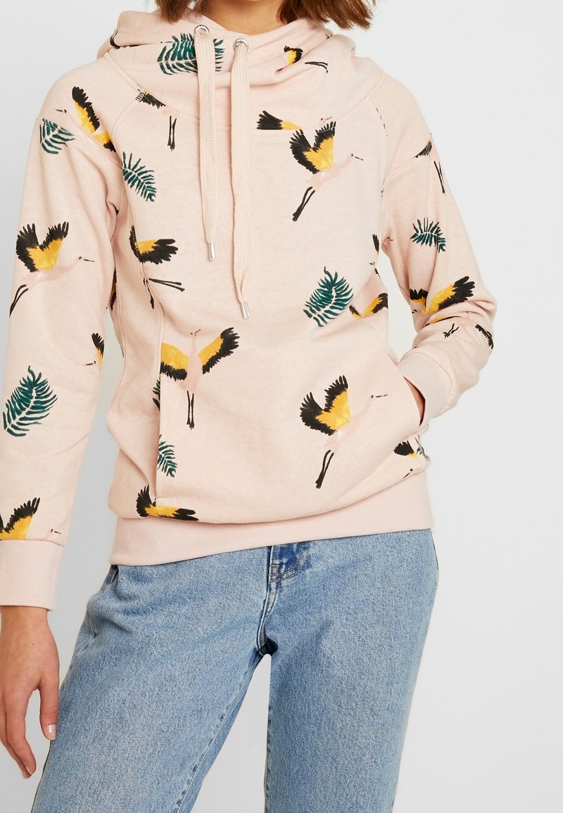 Femme portant un sweat à capuche rose clair avec des imprimés d'oiseau grue jaune et noir et des feuilles vertes, associé à un jean bleu clair.