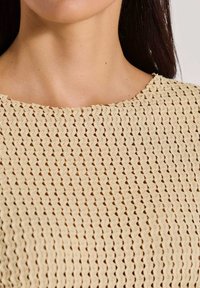 Vrouw in een beige gebreide top met een ronde halslijn en een gestructureerd, open weefpatroon, met steil, donker haar.