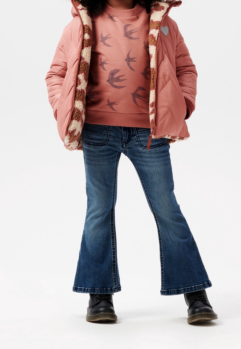 Enfant portant un pull rose à motif d’oiseaux, une doudoune rose avec doublure à motif, un jean évasé bleu et des bottes noires, debout les mains dans les poches.