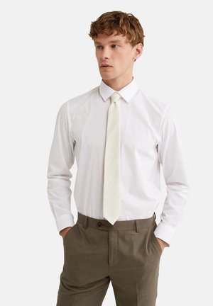 Camisa blanca de manga larga con cuello puntiagudo, combinada con una corbata color crema y pantalones marrones texturizados. Tejido suave con un corte ajustado.
