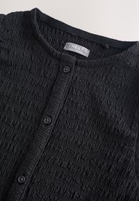 Chemise noire texturée boutonnée avec un col rond et trois boutons visibles, avec une étiquette "NEXT Made in Sri Lanka" à l'intérieur du col.