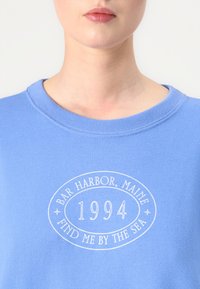 Svijetloplavi dukserica s okruglim motivom koji uključuje "Bar Harbor, Maine," "1994," i "Nađi me uz more" u bijelom tekstu. Mekana tkanina.