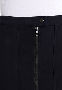 Svart kjol med en slät textur, utrustad med dragkedja fram och tryckknappsstängning. Utformad med vertikala sömmar för struktur.