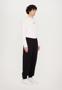 White long-sleeve shirt with black logo, paired with black sweatpants featuring elastic cuffs. White sneakers complete the outfit.