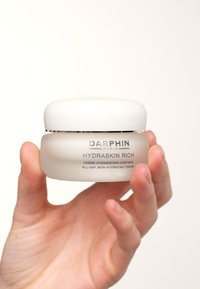 Witte glazen pot met een gladde, ronde deksel. Het etiket zegt "Darphin Hydraskin Rich" in grijze tekst. Een hand houdt de pot vast.