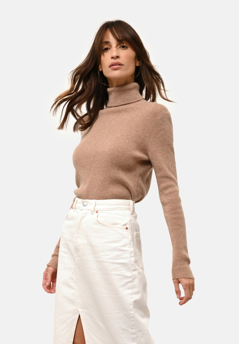 Beige gestreifter Rollkragensweater mit langen Ärmeln, kombiniert mit einem weißen Jeansrock, der einen Schlitz vorne und dezente Kupferhardware-Akzente aufweist.