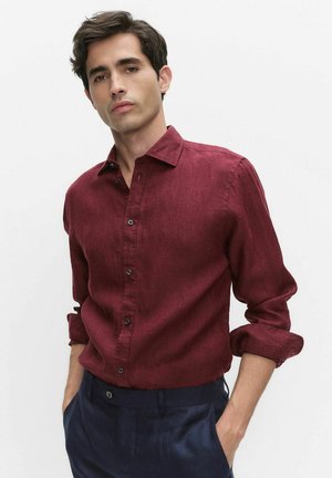 Mand iført en langærmet bordeaux skjorte med op rullede manchetter og marineblå bukser, stående med hænderne i lommerne.