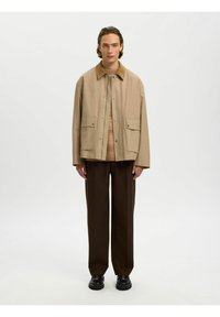 Homme debout portant une veste beige avec un col marron, un pull beige, un pantalon marron foncé et des chaussures noires sur fond blanc.