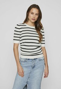 Suéter de punto a rayas en crema y negro con mangas cortas y cuello acanalado. Combinado con una falda de mezclilla clara con una abertura en la parte frontal.