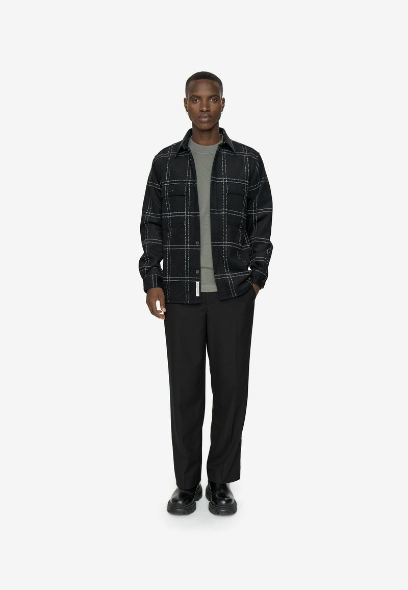 Sort ternet skjorte med knapper over en grøn top, parret med sorte bukser og kraftige sorte sko. Enkel og struktureret design.