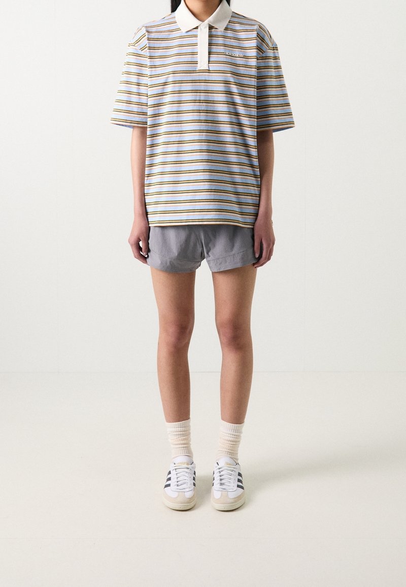 Personne portant un polo à manches courtes rayé, un short gris, des chaussettes claires et des baskets Adidas blanches avec des rayures noires, se tenant sur un sol blanc.