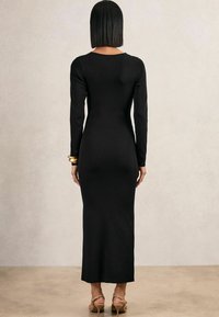 Persoană cu părul scurt negru poartă o rochie lungă neagră, strâmtă, brățară de aur la încheietura stângă și sandale bej cu toc înalt, stând pe un podea bej.