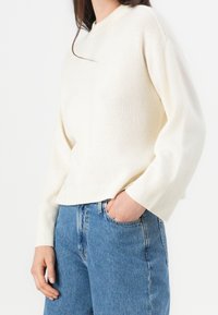Kremowy sweter z dzianiny z okrągłym dekoltem, ściągaczami na rękawach oraz luźnym krojem, zestawiony z jasnoniebieskimi jeansami.