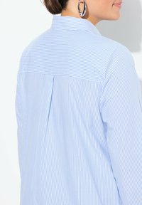 Camisa de rayas azul claro con un pliegue en la espalda, un cuello clásico y textura suave. Presenta un diseño de botón y sin hardware visible.