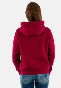 Sudadera con capucha de forro polar en rojo intenso, con capucha ajustable mediante cordón, puños y bajo acanalados. Combinada con vaqueros azules que presentan costuras decorativas en los bolsillos.