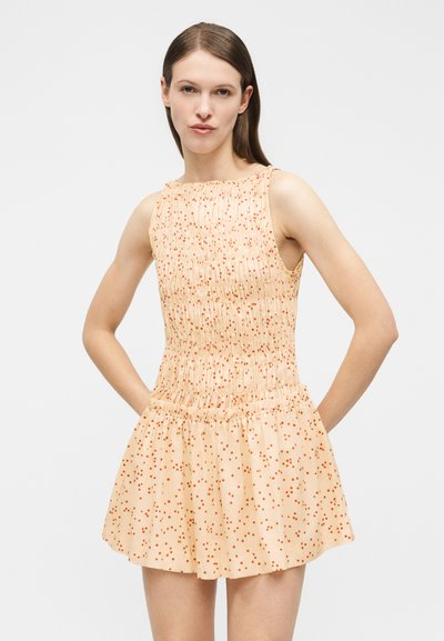 Γυναίκα που φοράει αμάνικο μπεζ φόρεμα με πορτοκαλί πουά, εφαρμόζον κορσάζ με smocking και φουσκωτή φούστα, στέκεται με τα χέρια στη μέση.