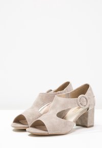 Scarpe open-toe in suede taupe con tacco basso a blocco, caratterizzate da tagli laterali e una fibbia rotonda decorativa sulla cinghia esterna alla caviglia.