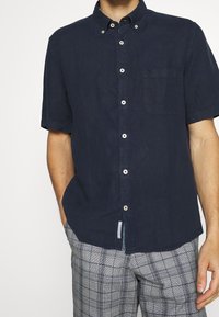 Marineblauw kortopgestoken overhemd van linnen, met een borstzak, witte knopen en een relaxed fit. Gepaard met geruite broek.