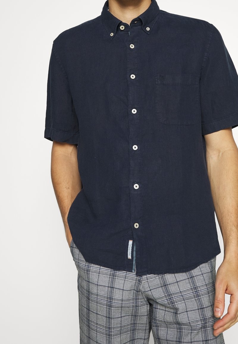 Marineblauw kortopgestoken overhemd van linnen, met een borstzak, witte knopen en een relaxed fit. Gepaard met geruite broek.