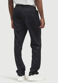 Schwarze Sporthose mit glatter Textur, elastischem Bund und einer Gesäßtasche; kombiniert mit weißen Turnschuhen.