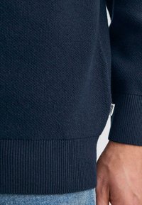 Maglione blu navy con tessuto testurizzato e orlo a costine. Etichetta visibile cucita di lato. Manica mostrata sopra un paio di jeans blu.