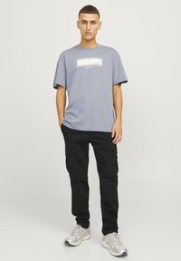 Camiseta de manga corta azul claro con un gráfico rectangular en beige y blanco; combinada con pantalones cargo negros y zapatillas plateadas.