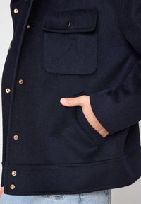 Giacca in misto lana blu navy con bottoni a pressione in tono oro, tasca sul petto e tasche laterali, caratterizzata da una trama morbida e una vestibilità rilassata.