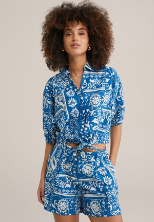 Camicetta corta a maniche corte con motivo blu, legata in vita. Abbinata a pantaloni corti coordinati, entrambi con motivi floreali bianchi.