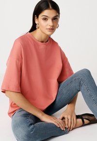 Koraalkleurig oversized t-shirt met korte mouwen, ronde halslijn, zachte textuur, gecombineerd met blauwe nauwsluitende jeans en zwarte platte schoenen.