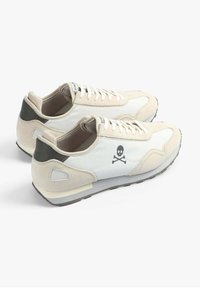 Baskets sportives en tissu crème avec des accents en daim. Elles disposent d'un logo noir de skull et os croisés, d'un détail de talon gris et d'une semelle en caoutchouc.