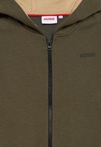 Hugo mikina na zip v olivově zelené barvě, s texturovanou tkaninou, černým zipem a subtilním logem na hrudi. Podšitá béžovou podšívkou.