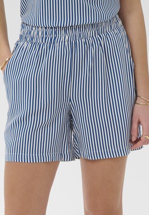 Vrouw draagt blauwe en witte verticale gestreepte shorts met elastische taille en zakken, handen ontspannen langs het lichaam, gouden armbanden om de polsen.