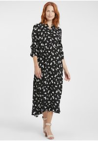 Schwarzes Midi-Kleid mit weißen Blumenmustern, langen Ärmeln, einem mit Knöpfen versehenen Kragen und gerafften Stufen. Getragen mit nacktfarbenen Riemchen-Heels.
